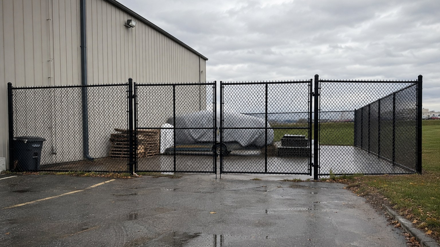 Clôture commerciale en mailles de chaîne noires, continue autour d'une cour industrielle au Québec, portail double battant fermé et verrouillé par une tige au sol