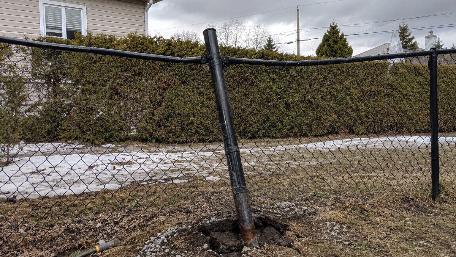 Single leaning steel post of a black chain-link fence with soil visibly heaved by frost at its base
