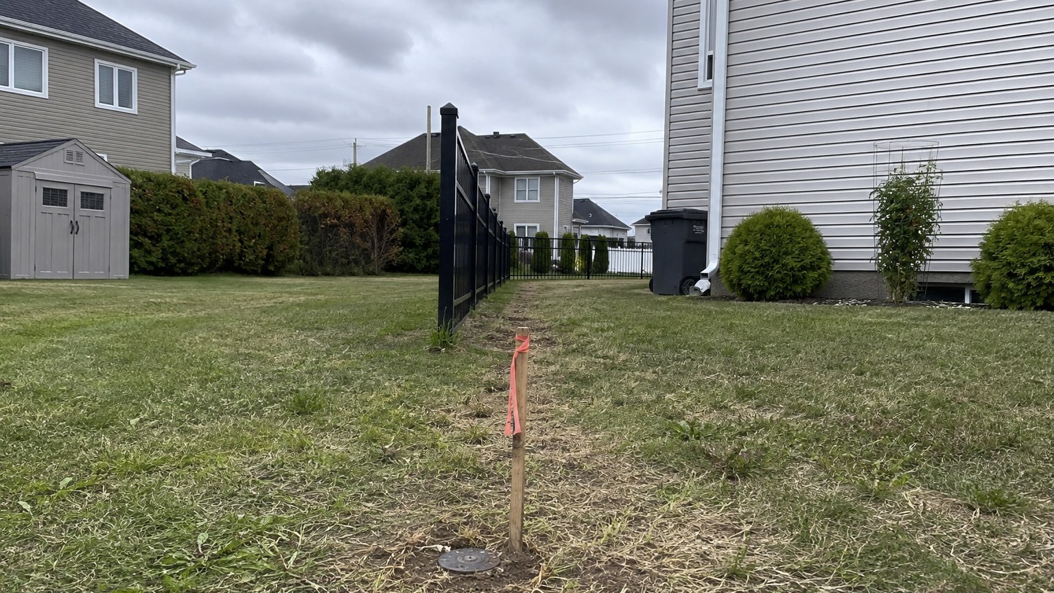 Iron survey pin with a flagged wooden stake marking the property line between two Quebec back yards, fence beginning beyond it